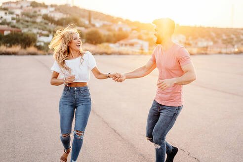 Fröhlicher Mann und aufgeregte Frau schauen sich an und lachen, während sie tanzend und Händchen haltend eine asphaltierte Straße entlang gehen, während die Stadt auf einem Hügel am Abend verschwimmt - ADSF39001