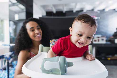 Happy baby boy playing on high chair with mother sitting in background - PNAF04537