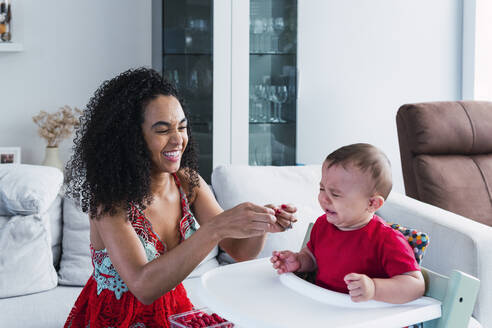 Happy mother with crying baby boy sitting in high chair at home - PNAF04540