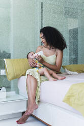 Curly-haired woman feeding baby boy with bottle on bed at home - PNAF04548
