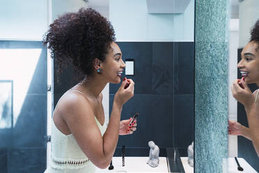 Happy woman with curly hair applying lipstick in bathroom at home - PNAF04557