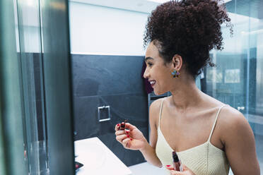 Happy woman applying lipstick standing in bathroom at home - PNAF04560