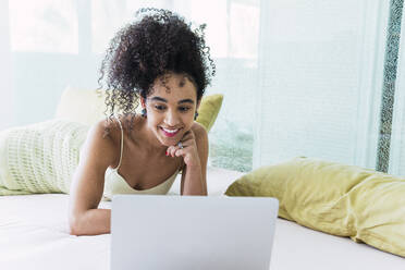 Happy woman with curly hair using laptop on bed at home - PNAF04566
