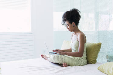 Curly-haired woman using laptop sitting on bed at home - PNAF04567