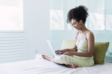 Smiling woman with curly hair using laptop sitting on bed at home - PNAF04568