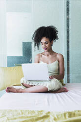 Lockenköpfige Frau benutzt Laptop auf dem Bett zu Hause - PNAF04569