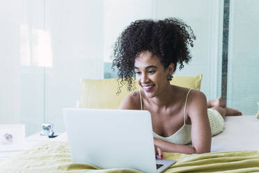 Glückliche Frau mit lockigem Haar liegt auf dem Bett und benutzt einen Laptop zu Hause - PNAF04571