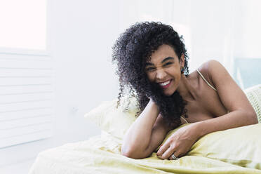 Cheerful curly-haired woman lying on bed at home - PNAF04578