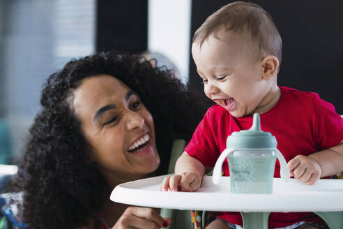 Cheerful mother playing with baby boy sitting on high chair at home - PNAF04582