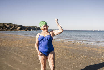 Senior woman in swimsuit showing fists at beach on sunny day - UUF27323
