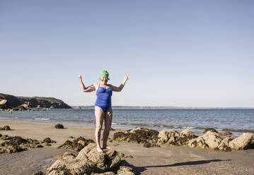 Ältere Frau steht auf einem Felsen und lässt Muskeln am Strand spielen - UUF27344