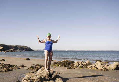 Ältere Frau steht auf einem Felsen und lässt Muskeln am Strand spielen - UUF27344