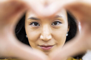 Smiling woman looking through hands making heart shape - WPEF06492