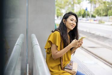 Lächelnde Frau mit Mobiltelefon an der Straßenbahnhaltestelle - WPEF06495
