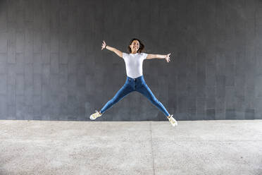 Happy woman with arms outstretched jumping on footpath in front of gray wall - WPEF06501