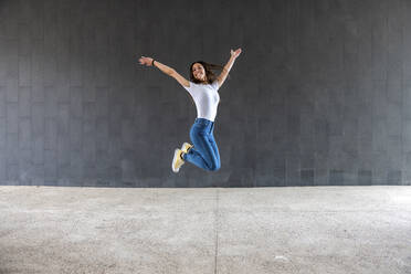 Happy woman jumping on footpath in front of gray wall - WPEF06503