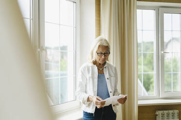 Frau hält Zeitung und liest in der Nähe des Fensters zu Hause - SEAF01255