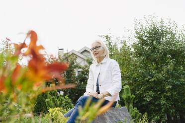 Thoughtful woman sitting on rock amidst plants in garden - SEAF01271