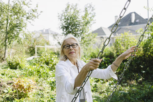 Senior woman holding chain of swing in garden - SEAF01292