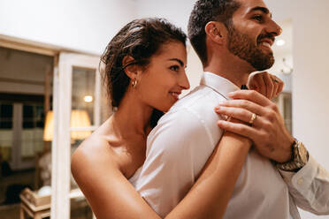 Woman fixing her husband's bow tie in a hotel . Happy young woman smiling while standing behind her husband in a white towel. Romantic young couple dressing up for a date in in a luxury hotel. - JLPPF00993