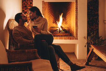 Flitterwöchner entspannen sich vor einem Kamin in einem Hotel. Romantische junge Frau sitzt auf dem Schoß ihres Mannes und hält ein Glas Wein in der Hand. Junges Paar genießt seine Flitterwochen in einem Luxushotel. - JLPPF01008