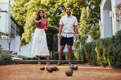 Happy young tourist couple playing a game of petanque together outdoors at a hotel. Young couple smiling cheerfully while spending quality time together. Couple bonding during a vacation. - JLPPF01015