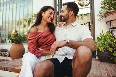 Young couple smiling at each other while sitting outside a luxury hotel. Romantic young couple enjoying their honeymoon vacation. Tourist couple bonding at a holiday resort. - JLPPF01022