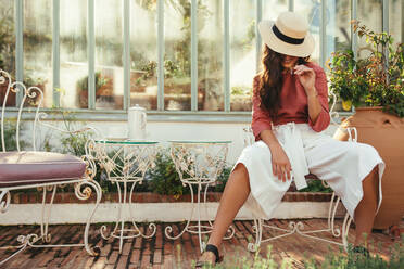 Fröhliche junge Touristin lächelt, während sie alleine vor einem Luxushotel sitzt. Modische junge Frau sitzt mit einem Zipfelhut während des Tages. Frau, die alleine an einem Urlaubsort Urlaub macht. - JLPPF01036