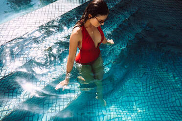 Young woman walking into a swimming pool while wearing a swimsuit. Attractive young tourist woman going for a swim at a holiday resort. Woman vacationing alone at a luxury retreat. - JLPPF01050