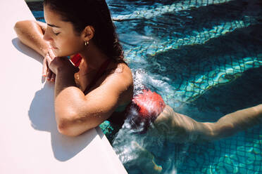 Young tourist woman relaxing in a swimming pool during the day. Attractive young woman leaning on the edge of a swimming pool with her eyes closed. Woman unwinding on a summer vacation. - JLPPF01052