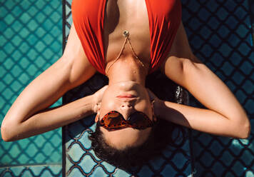 Relaxing in the sun during summer. Attractive young tourist woman sunbathing by the swimming pool at a luxury spa resort. Young woman vacationing alone at a holiday retreat. - JLPPF01064