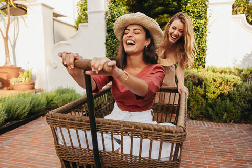 Female friends having fun with a trolley cart outdoors. Cheerful young woman pushing her friend in a trolley cart at a holiday resort. Two happy young women enjoying their summer vacation together. - JLPPF01075