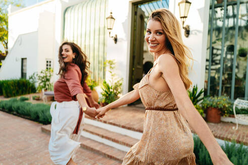 Woman leading her friend by the hand outside a holiday resort. Two happy young women smiling cheerfully while running together. Best friends having fun on their summer vacation. - JLPPF01098