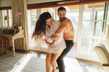 Carefree couple being playful in a hotel. Happy young woman having fun wearing her husband’s shirt in the morning. Romantic young couple enjoying their honeymoon getaway in a luxury hotel. - JLPPF01141