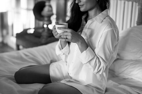 Brünette Frau genießt eine Tasse Kaffee in ihrem Hotelzimmer. Attraktive junge Touristin im weißen Hemd auf ihrem Bett sitzend. Junge Frau genießt einen Wochenendausflug in einem Luxushotel. - JLPPF01169