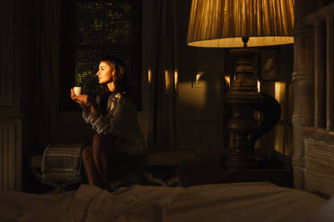 Gelassene junge Frau, die sich in ihrem Hotelzimmer entspannt. Attraktive junge brünette Frau, die eine Tasse Kaffee im Dunkeln trinkt. Junge Touristin, die einen Wochenendausflug in einem Luxushotel genießt. - JLPPF01181
