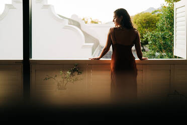Tourist woman standing outside her hotel room. Rearview of an attractive young woman looking over her shoulder in a red dress. Young woman vacationing alone in a luxury hotel. - JLPPF01185