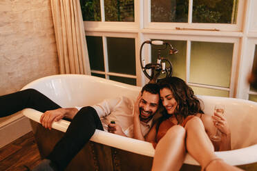 Fröhliche junge Frau umarmt ihren Mann, während sie in einer Badewanne sitzt. Romantisches junges Paar trinkt Champagner während ihrer Flitterwochen. - JLPPF01193