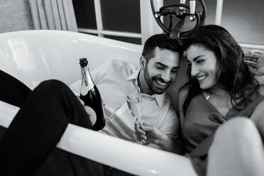 Fröhliches junges Paar, das in einer Hotelbadewanne mit Champagner anstößt. Romantisches junges Paar, das während seiner Flitterwochen in einem Luxushotel feiert. - JLPPF01197
