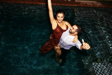 Flitterwöchner, die jubelnd in einem Swimmingpool stehen. Blick von oben auf ein glückliches junges Paar, das sich nachts in einem Pool vergnügt. Romantisches junges Paar, das seinen Flitterwochenurlaub in einem Luxushotel genießt. - JLPPF01205