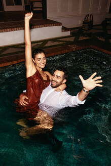 Couple smiling and cheering in a swimming pool. Happy young couple having fun together in a swimming pool at night. Romantic young couple enjoying their honeymoon vacation at a luxury hotel. - JLPPF01208