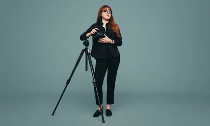 Pensive photographer holding a digital camera while standing next to her tripod. Young professional photographer looking away thoughtfully while standing against a studio background. - JLPPF01266