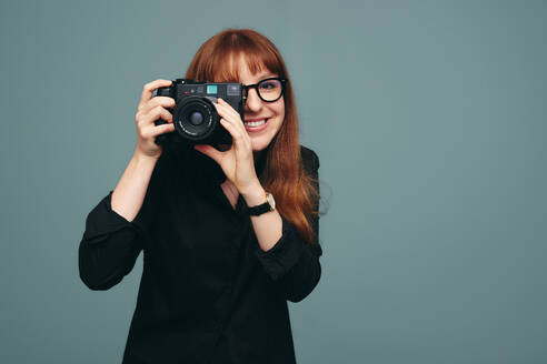 Happy female photographer taking a picture on her digital camera. Young professional photographer looking through camera lenses while standing against a studio background. - JLPPF01269