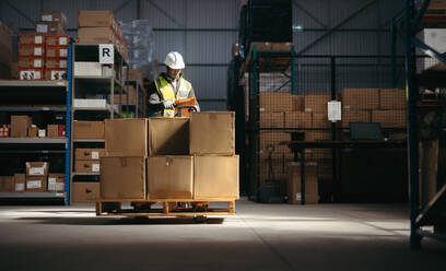 Logistics worker reading a clipboard while standing behind a pallet jack loaded with cardboard boxes. Man in a reflective jacket taking packages for storage in a distribution warehouse. - JLPPF01305