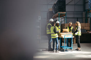 Fröhlich lächelnde Gruppe von Lagerarbeitern bei einer Besprechung mit ihrem Vorgesetzten. Gut gelaunte Logistikmitarbeiter, die in einem großen Vertriebszentrum als Team arbeiten. - JLPPF01306