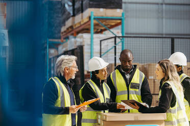 Diverse warehouse workers discussing some reports during a staff meeting. Group of multicultural logistics employees working as a team in a large distribution warehouse. - JLPPF01307