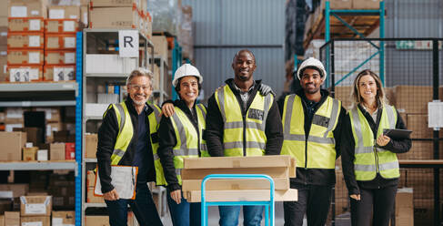 Porträt von glücklichen Lagerarbeitern, die in Arbeitskleidung zusammenstehen und in die Kamera lächeln. Gruppe von multikulturellen Logistikmitarbeitern, die in einem großen Vertriebszentrum als Team arbeiten. - JLPPF01308