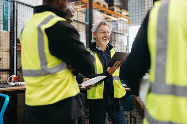 Älterer Lagerleiter, der bei einer Besprechung mit seinem Team fröhlich lächelt. Gruppe multikultureller Logistikmitarbeiter, die in einem großen Vertriebslager zusammenarbeiten. - JLPPF01309