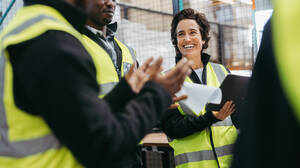 Ältere Frau, die während einer Besprechung mit ihren Kollegen in einem Auslieferungslager fröhlich lächelt. Gruppe von multikulturellen Mitarbeitern, die in einem großen Logistikzentrum zusammenarbeiten. - JLPPF01311
