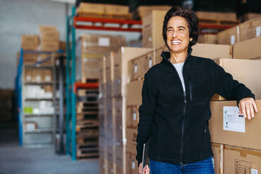 Glücklicher Logistikmanager, der lächelnd neben einem Stapel Kartons steht. Fröhliche reife Frau, die in einem großen Auslieferungslager arbeitet. - JLPPF01316